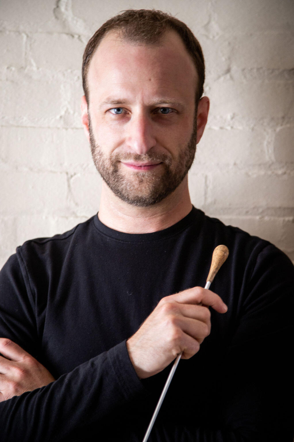 Photo by Wendy Redfern Dr. Matthew Mainella looks at the camera with a small, relaxed smile. The lighting is dramatic, highlighting the left side of his face and casting a shadow on the right side. His conducting baton is held loosely in one hand across his chest.