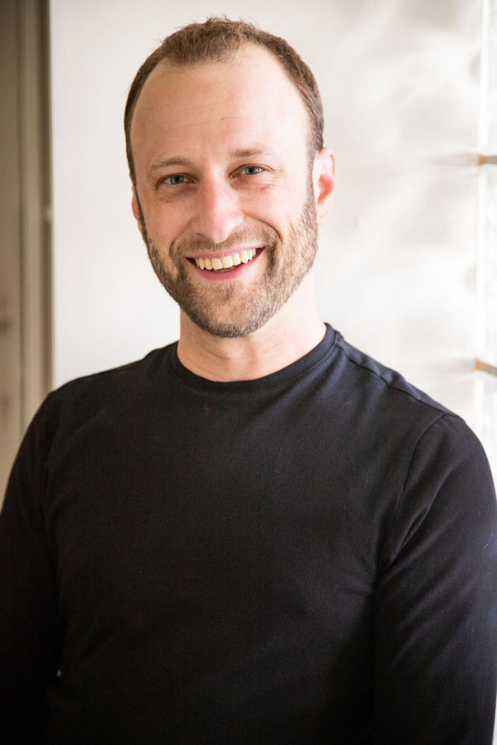 Photo by Wendy Redfern Dr. Matthew Mainella smiles at the camera, his head tilted to the side, his eyes focused on the viewer. He wears casual concert black and is leaning in front of a white-washed wall.